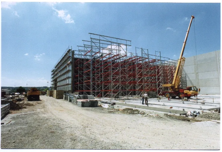 Eine Baustelle mit einem großen Stahlgerüst, einem gelben Kran, zwei Arbeitern, Betonplatten und verstreuten Baumaterialien unter einem teilweise bewölkten Himmel.