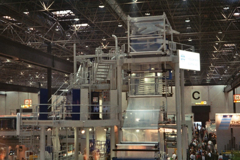 Eine große Industriemaschine, die in einer Messehalle transparente Kunststofffolie verarbeitet, im Hintergrund sind Menschen zu sehen, und darüber erstrecken sich hohe Decken mit Metallkonstruktionen.