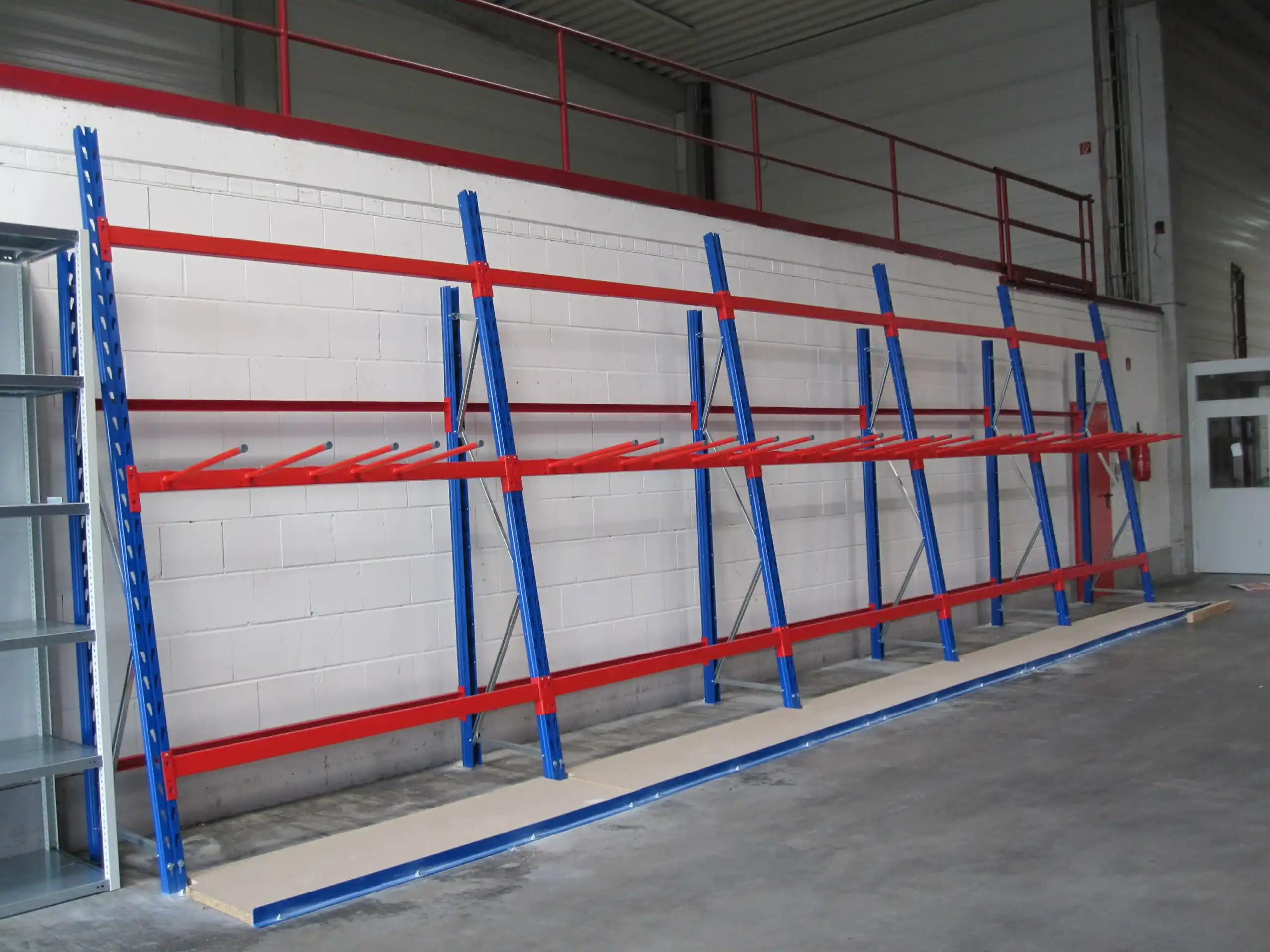 In einem Lagerraum steht ein blau-rotes Kragarmregal aus Metall vor einer weißen Backsteinwand; es verfügt über mehrere Arme zur Lagerung langer Gegenstände. Der Boden besteht aus Beton, und auf einem Zwischengeschoss darüber ist ein Metallgeländer zu sehen.