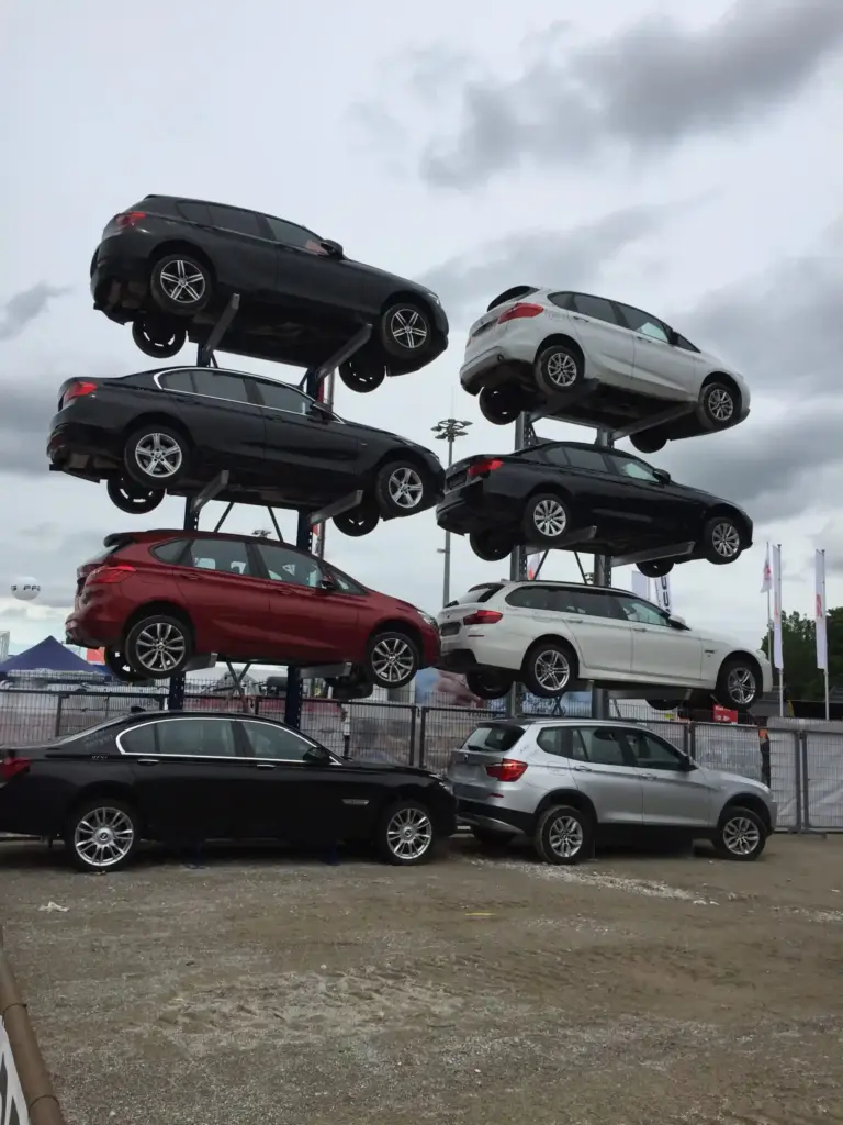 Sechs Autos sind in zwei vertikalen Dreierreihen übereinander gestapelt und balancieren über zwei weiteren, auf dem Boden geparkten Autos – alles innerhalb eines umzäunten Bereichs unter bewölktem Himmel.