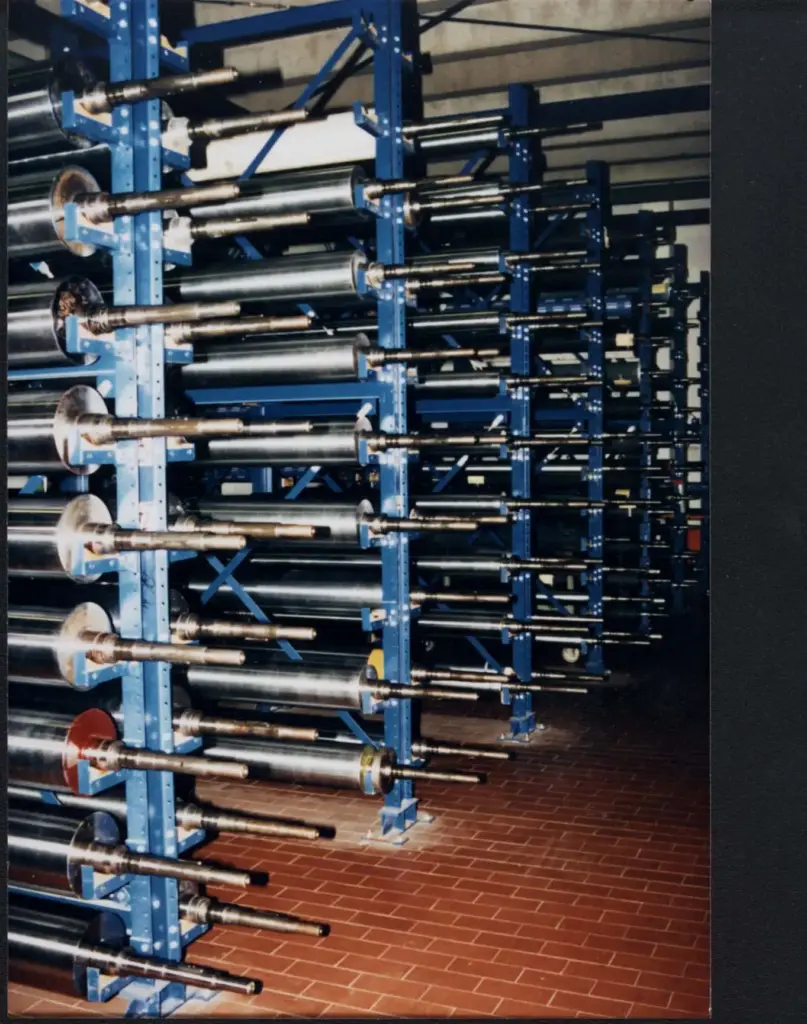 Reihen großer Metallspulen, die auf blauen Industrieregalen in einer Fabrikumgebung montiert sind, mit Fliesenboden und einer übersichtlichen Anordnung, was auf eine Produktions- oder Verarbeitungsstätte hindeutet.