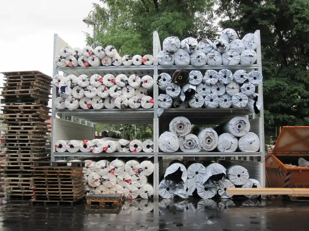 In Plastikfolie eingewickelte Materialrollen lagern im Freien auf Metallregalen, neben gestapelten Holzpaletten und einem Müllcontainer, im Hintergrund sind Bäume zu sehen.