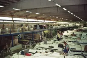 Eine große Werkshalle, in der Arbeiter unter greller Industriebeleuchtung weiße Bootsrümpfe zusammenbauen und bearbeiten, umgeben von Maschinen und Materialien.