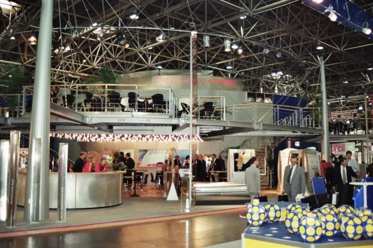 Eine belebte Messe- oder Ausstellungshalle, in der Menschen umhergehen und an verschiedenen Ständen miteinander interagieren, mit modernen Metallkonstruktionen, einem Sitzbereich im Obergeschoss und heller Beleuchtung von oben.
